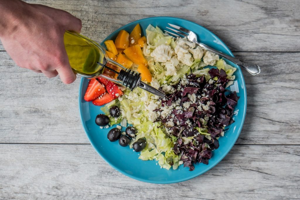 cuisine végétarienne : salade verte avec chou fleur cru, melon, fraise, myrtille, olives noires. Main qui verse de l'huile dessus